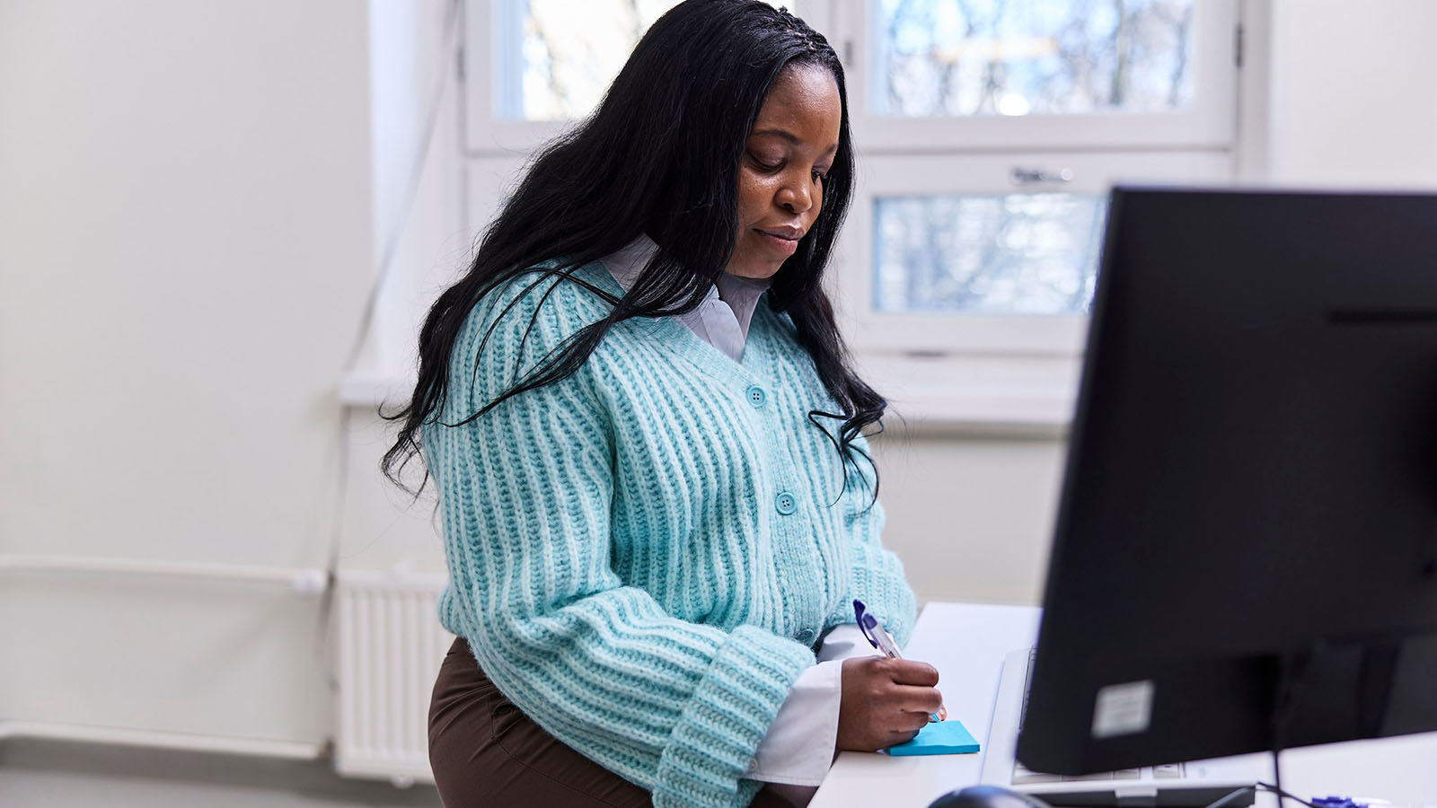 Terveydenhuollossa työskentelevä nainen istuu tietokoneen ääressä ja kirjoittaa jotain lapulle.